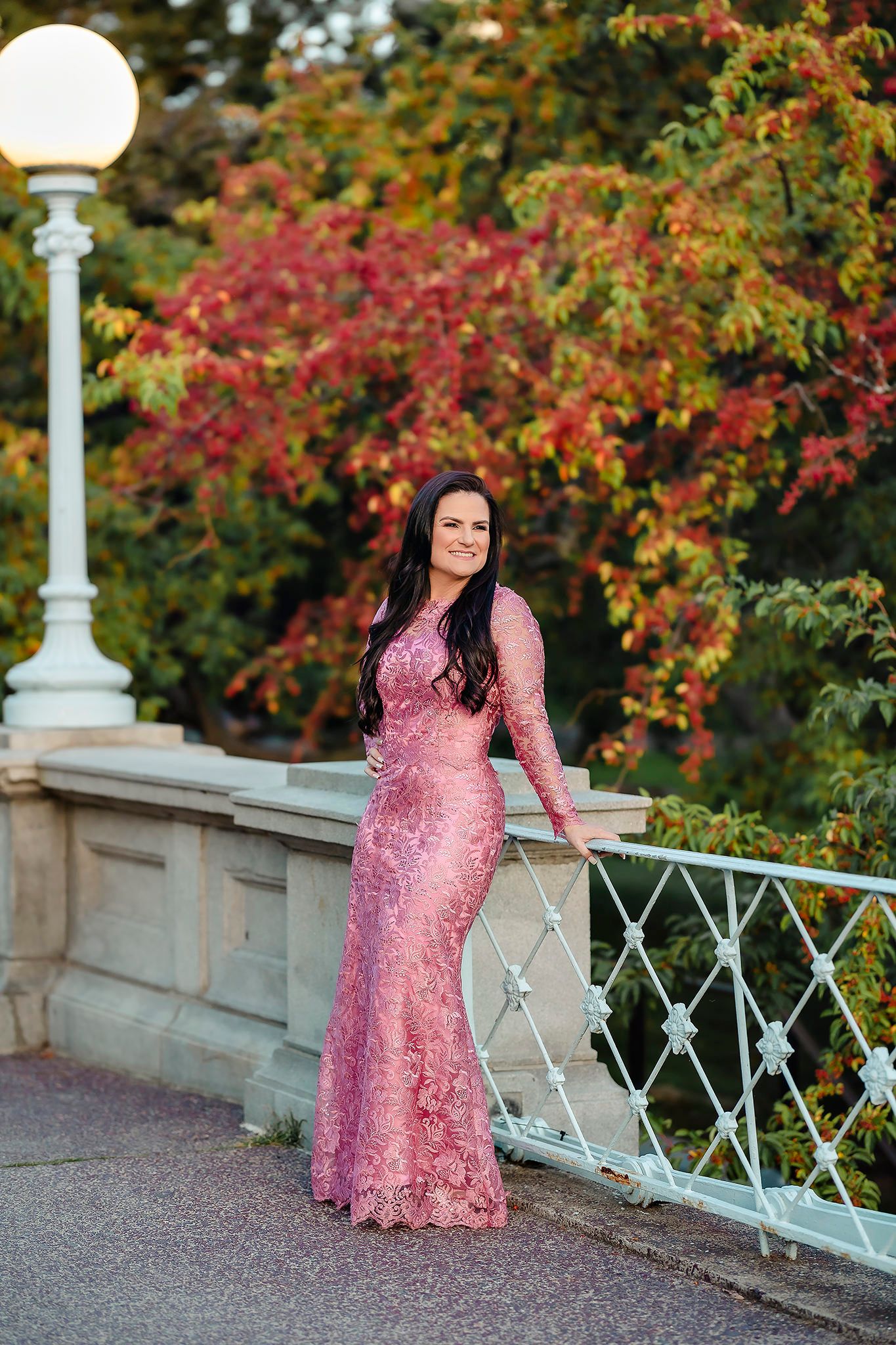 Woman in pink lace gown poses on bridge with autumn foliage backdrop.
