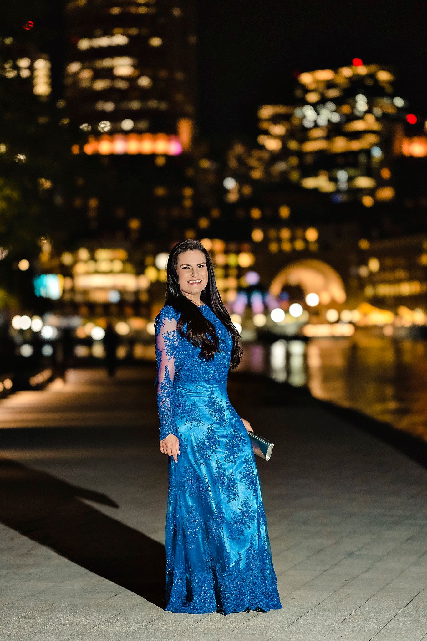 Woman in blue evening gown poses against cityscape at night.