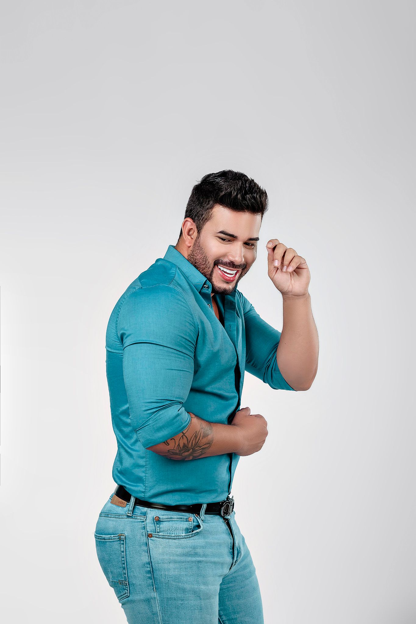 Model wearing teal shirt and jeans poses expressively against light background.
