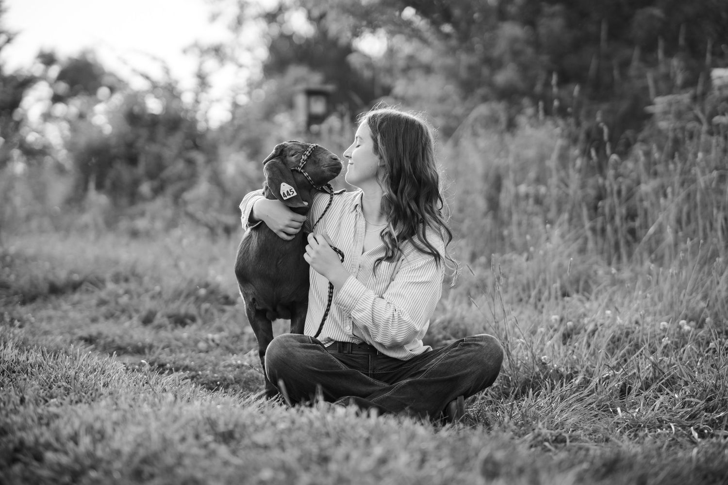 Black and white photo of a young woman sitting in grass, affectionately holding her market goat outdoors.