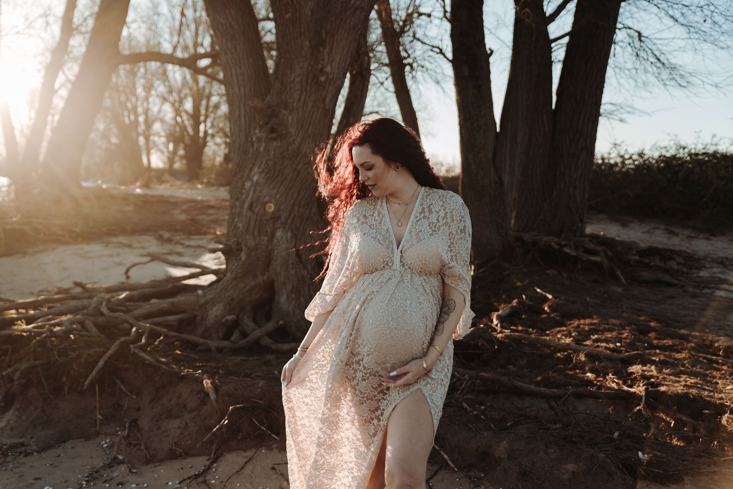 Sun-drenched maternity photoshoot in a white lace dress among bare winter trees.