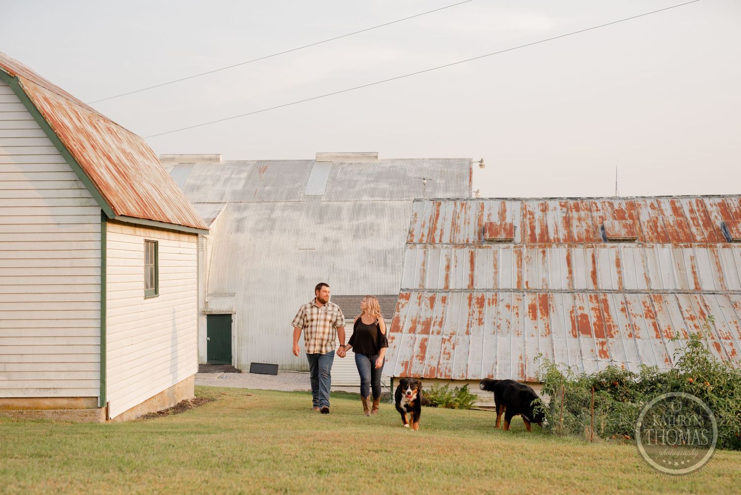 A couple walks with their dogs alongside a rustic barn with weathered metal roofing.