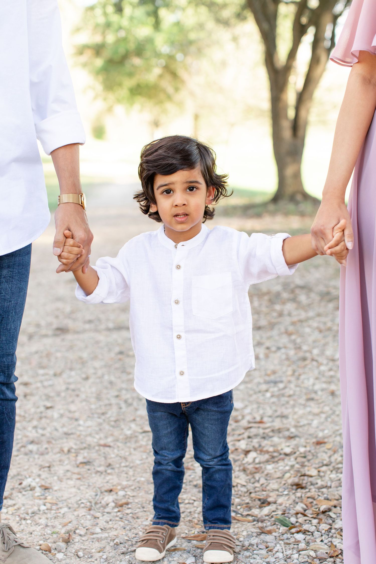 A small child in a white button-down shirt holds hands with parents on a tree-lined path.