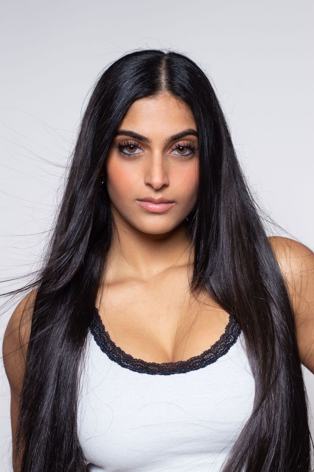 Beauty portrait against white backdrop showing long dark hair and white top with lace trim.