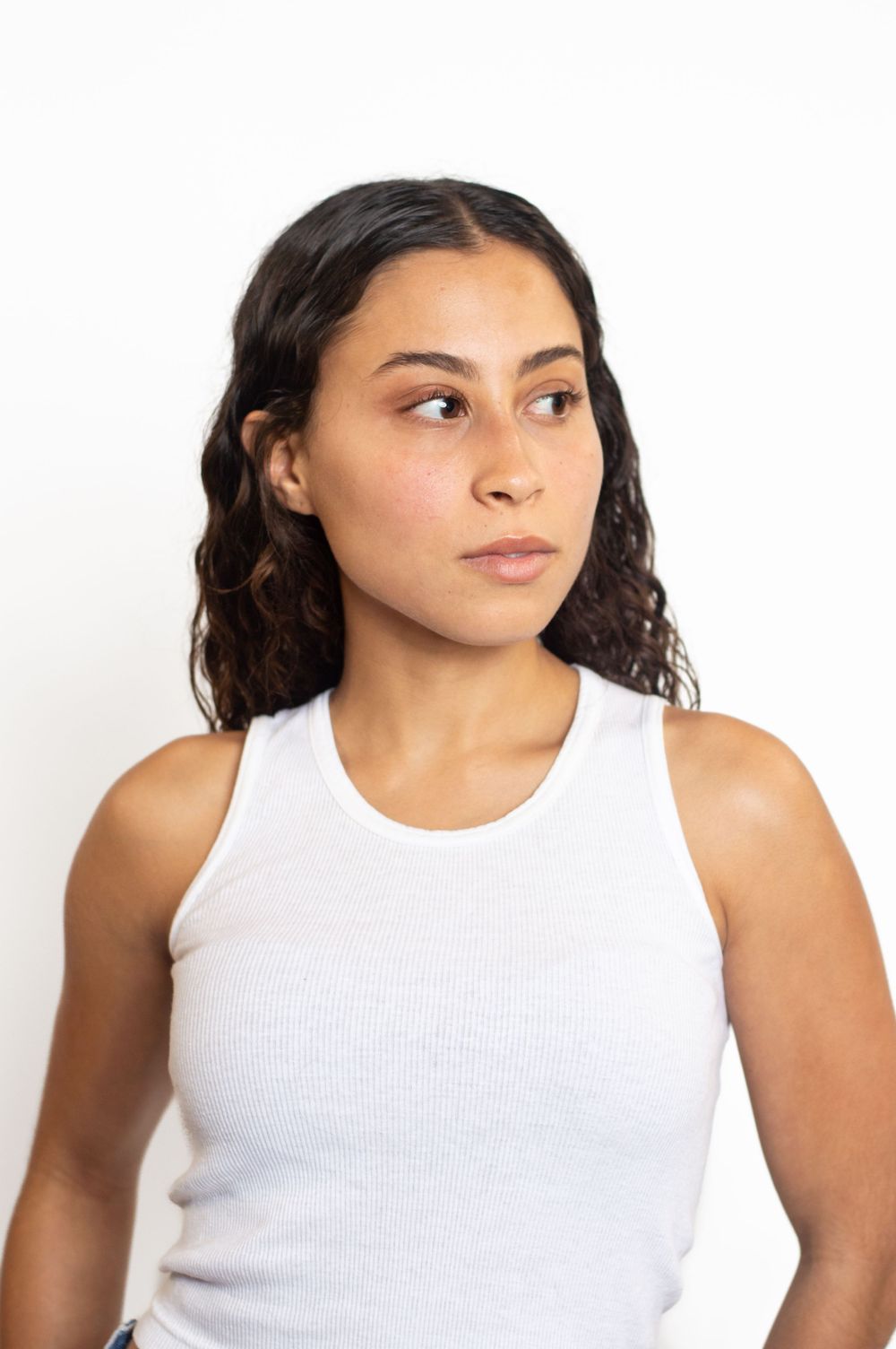 Portrait against white background featuring casual white tank top and natural wavy hair.