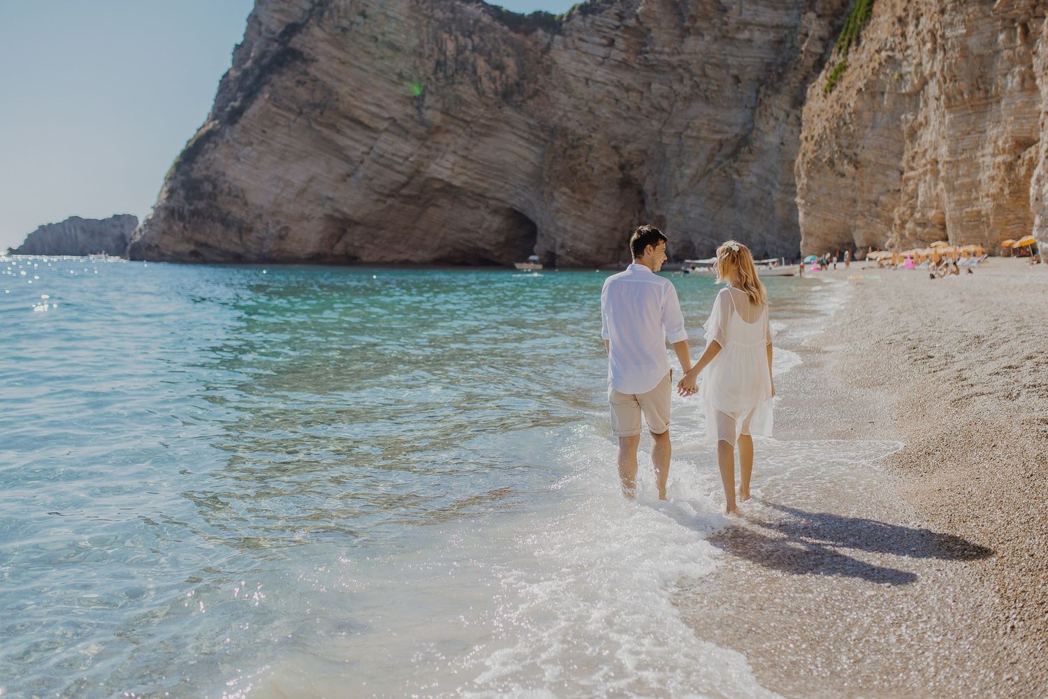 Couple walking along a scenic Mediterranean beach with crystal clear turquoise waters and limestone cliffs.