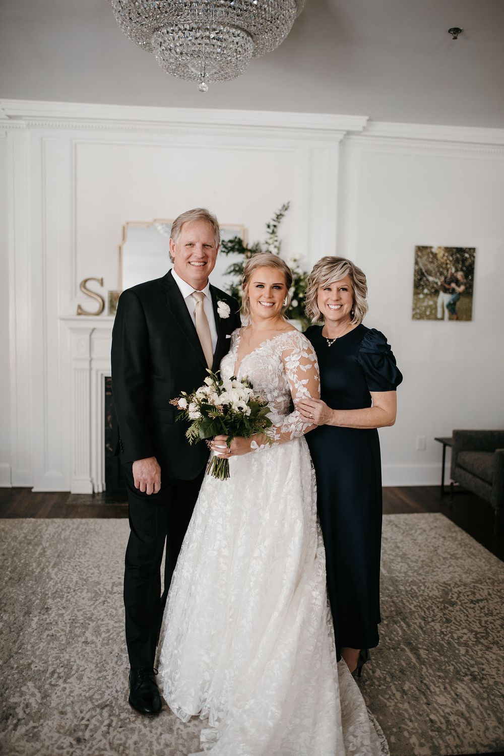 Bride in lace gown holds white bouquet while posing with her parents inside an elegant wedding venue.