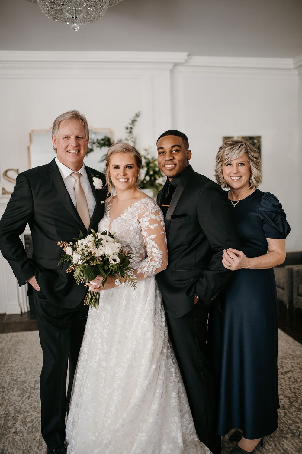 Bride and groom pose with her parents under a crystal chandelier inside a elegant white wedding venue.