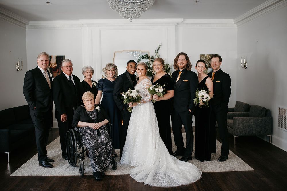 Large wedding family portrait gathered inside an elegant white venue with crystal chandelier overhead.