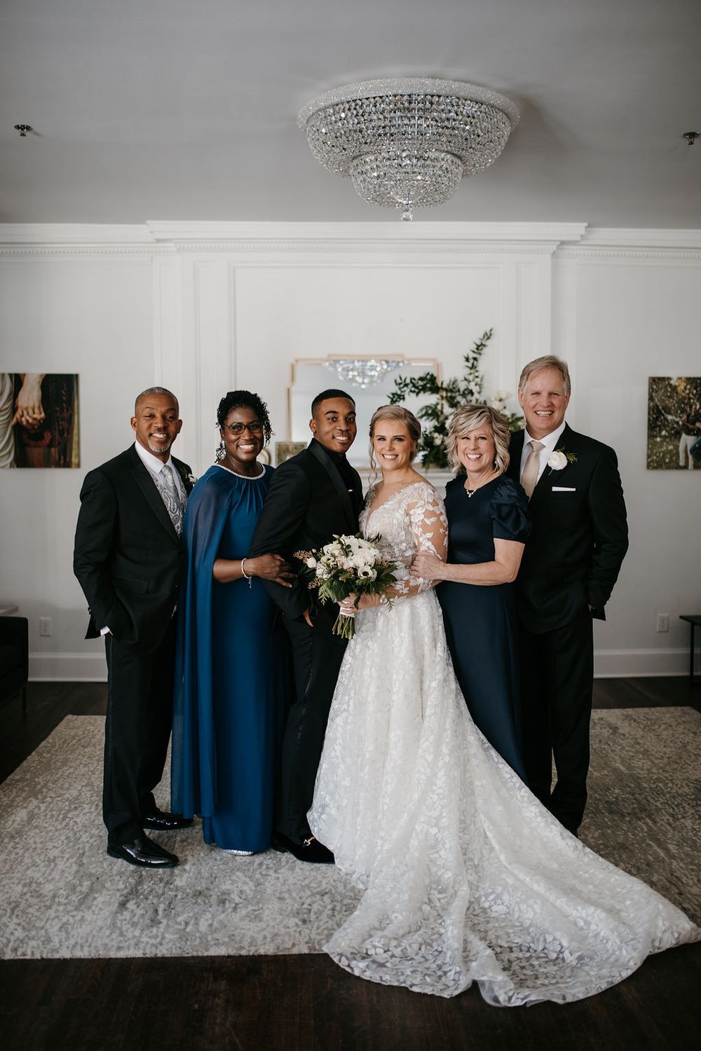 Bride and groom pose with both sets of parents in an elegant white room with a crystal chandelier.
