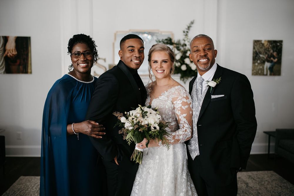 Bride and groom smile with grooms parents inside a bright elegant wedding venue with floral backdrop.