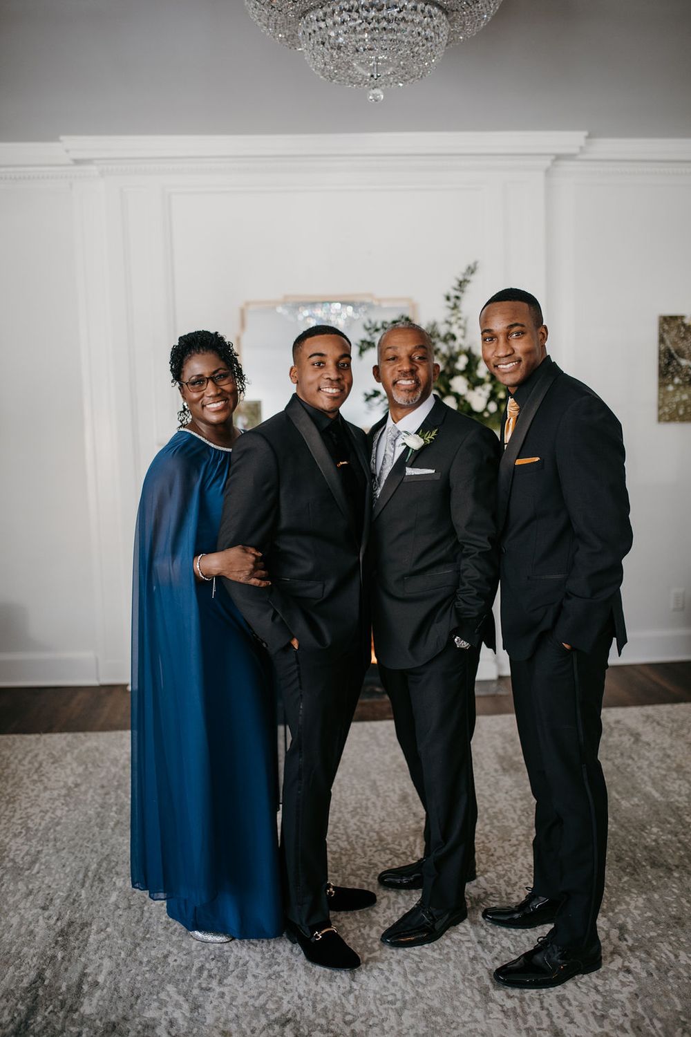 Groom poses with his mother in blue gown, father, and brother in black tuxedos at wedding venue.