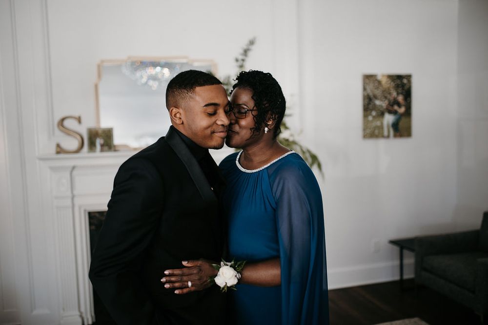 Groom in black tuxedo shares a tender forehead kiss with his mother in a blue gown at the wedding.