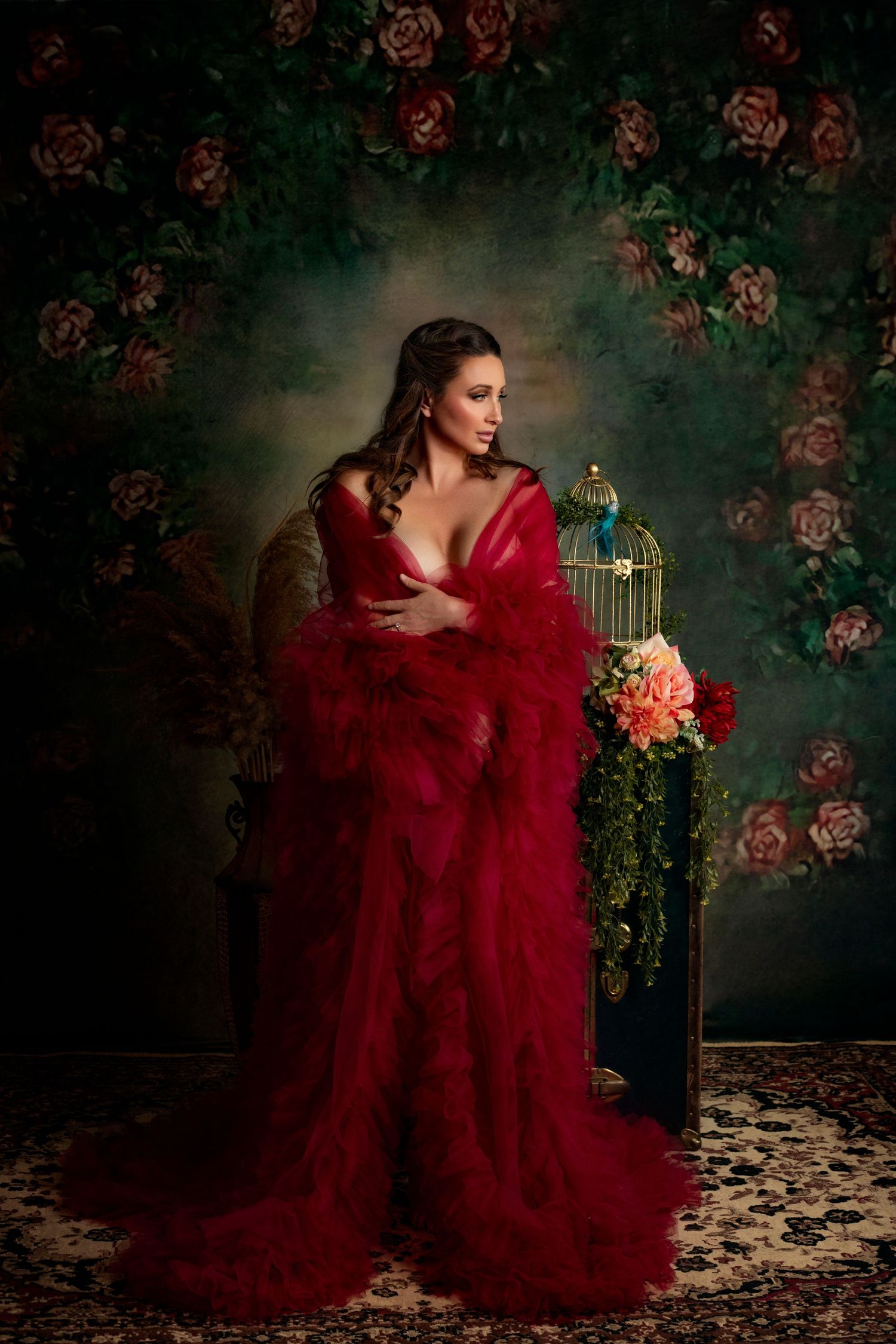 Dramatic portrait in a flowing red gown against a moody floral backdrop with vintage birdcage.