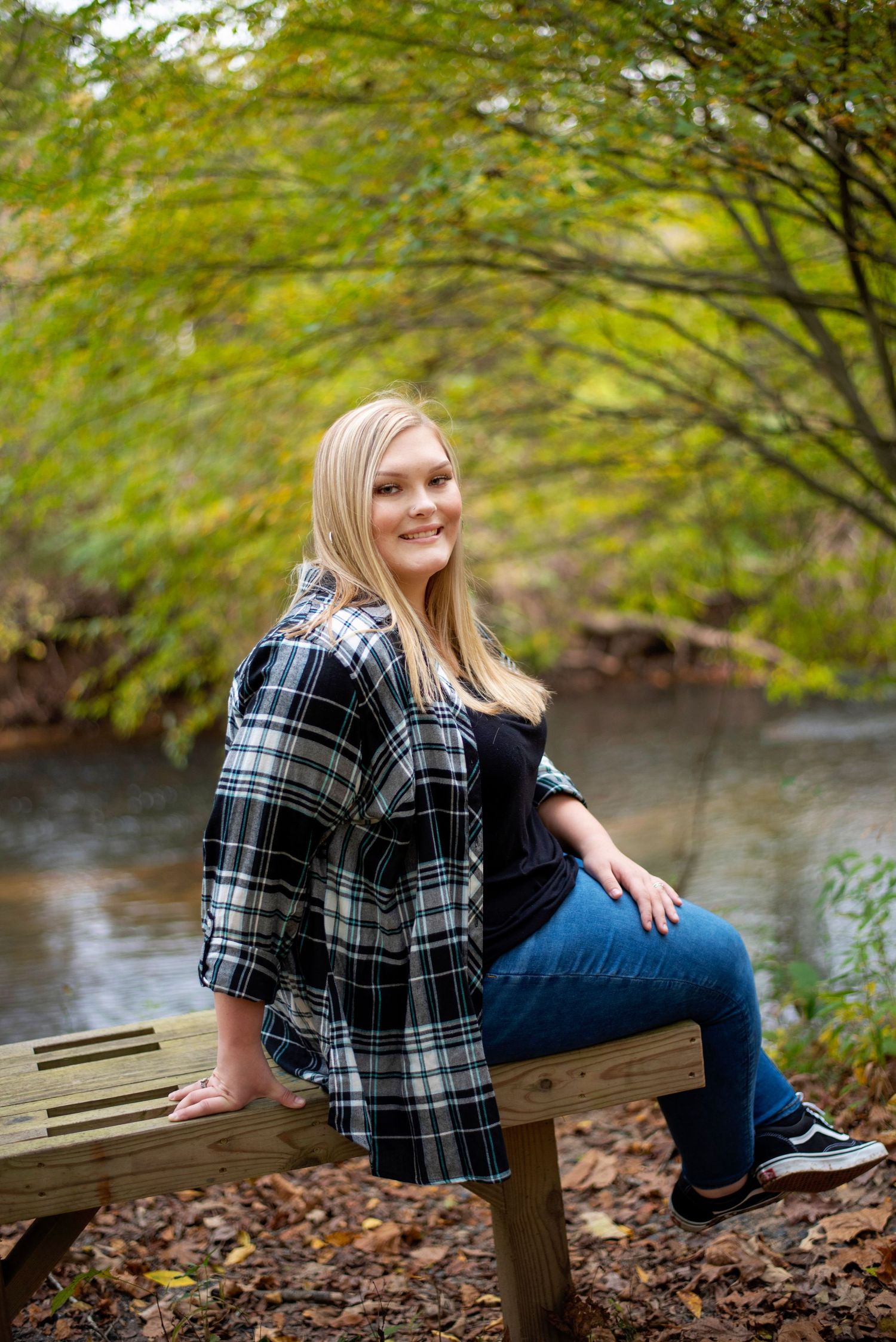 Woman in plaid shirt and jeans sits on wooden bench by autumn stream.