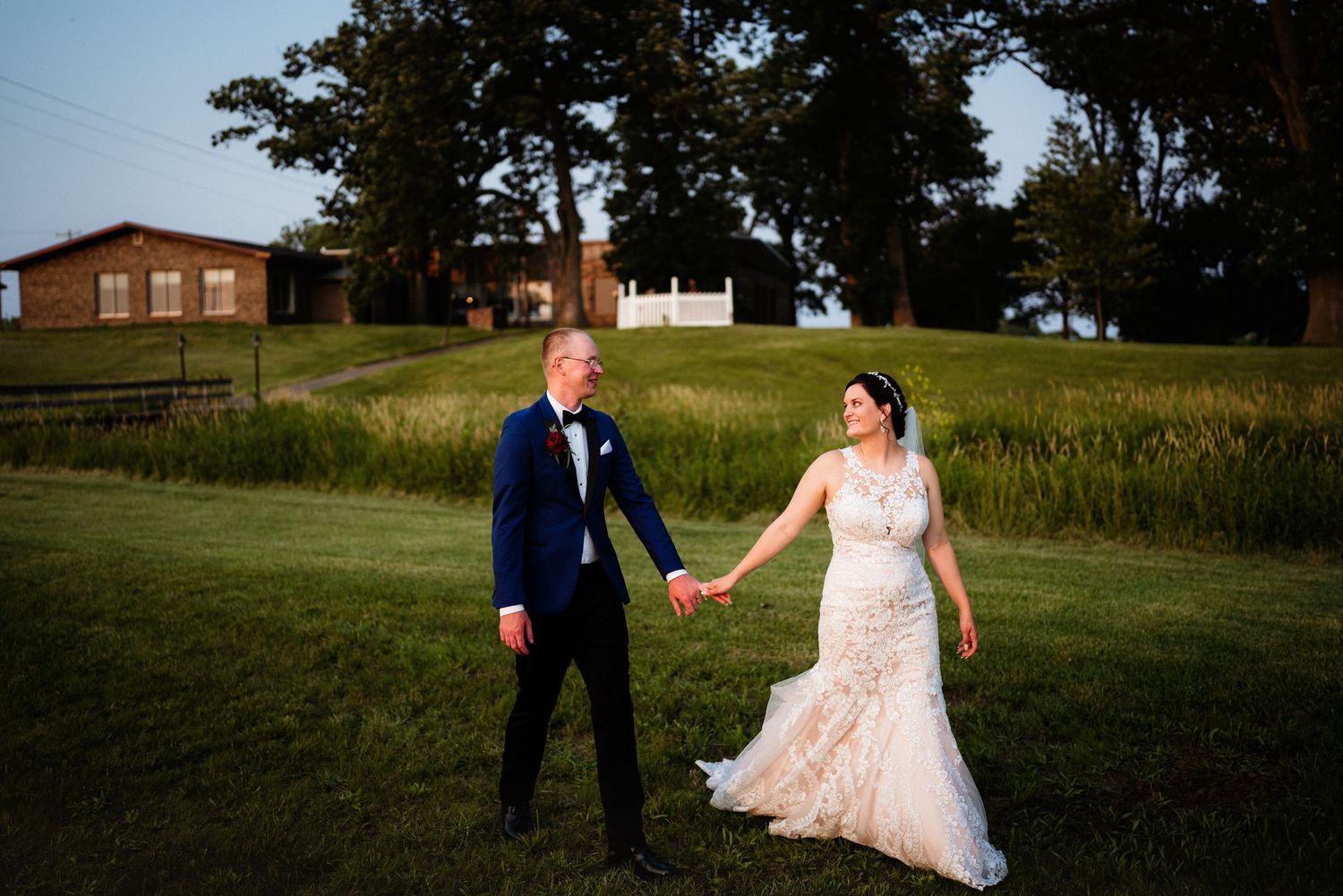 A couple in wedding attire holds hands on a grassy hill with a rustic barn in the background during sunset.