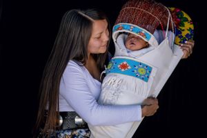 Native American woman holding infant in ornate beaded cradleboard against dark background.