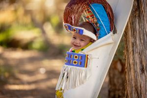 Smiling Native American baby in traditional beaded cradleboard with colorful regalia outdoors.