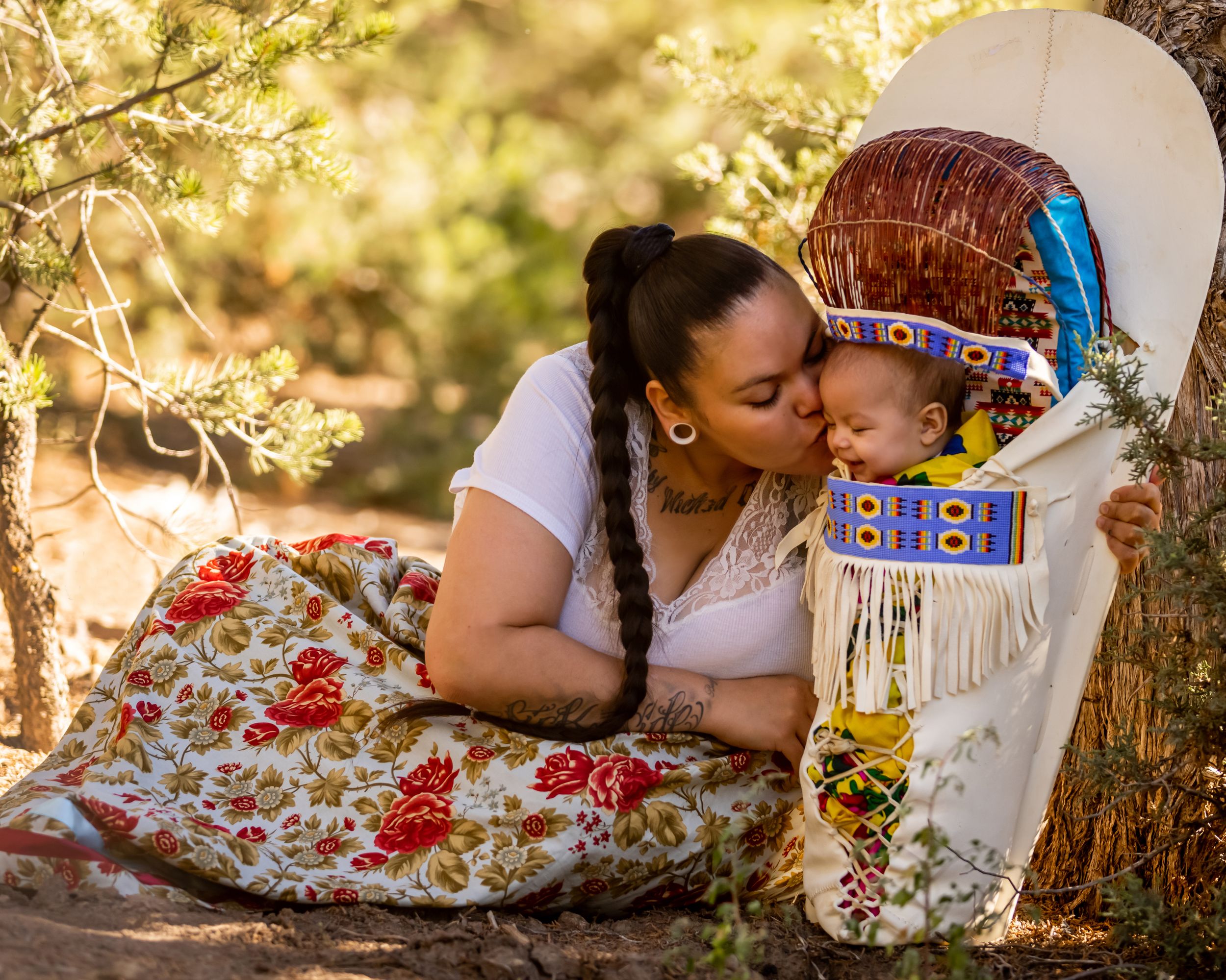 Native American mother kissing baby in traditional beaded cradleboard among trees.