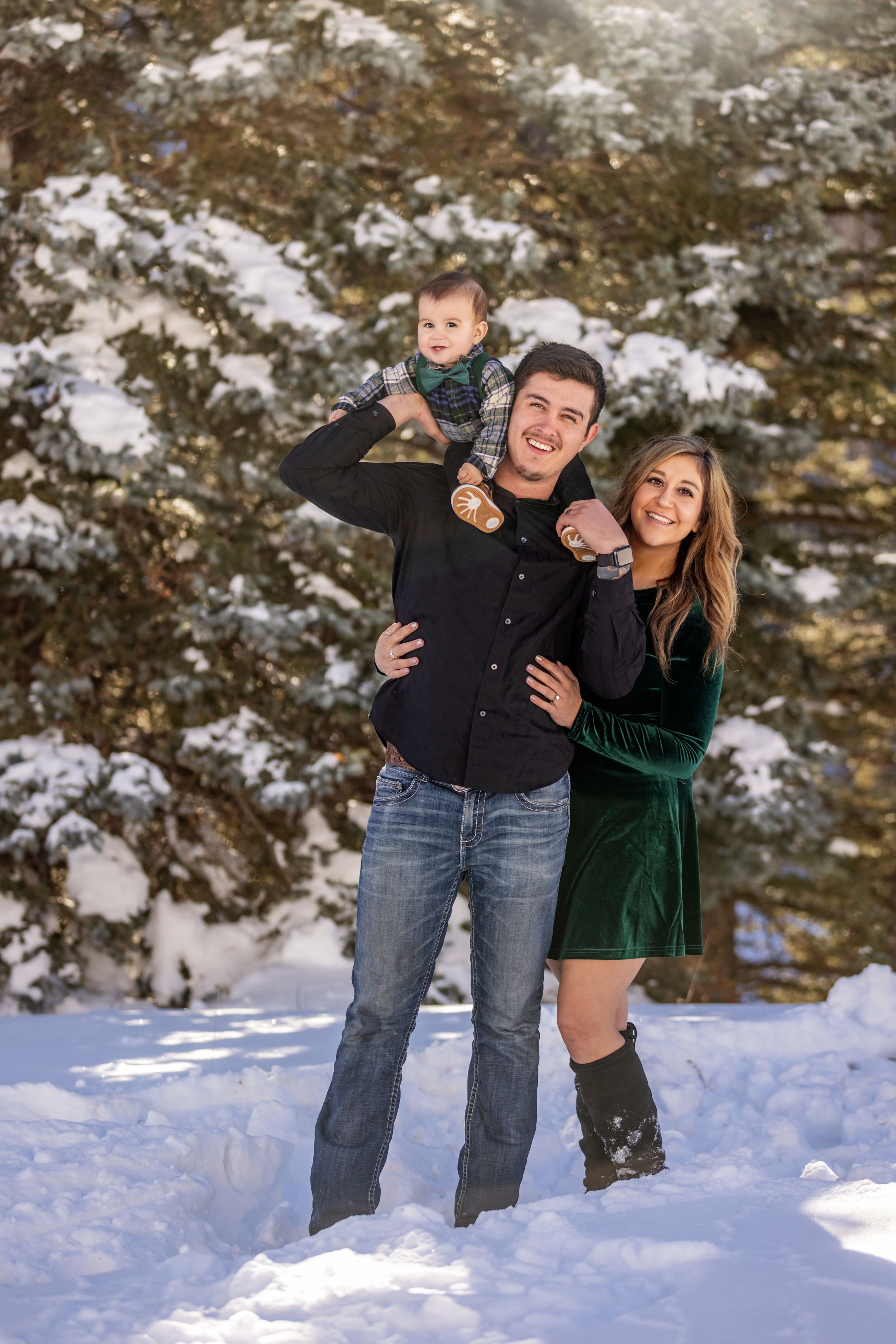 Smiling couple with baby in snowy winter landscape surrounded by snow-covered pine trees.