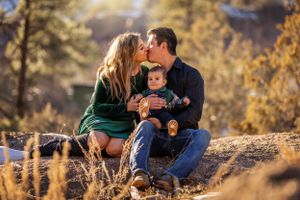 Couple kissing while holding their baby on a rocky outdoor setting in warm sunlight.