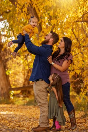 Family of four in golden autumn park, father lifting baby girl joyfully in the air.