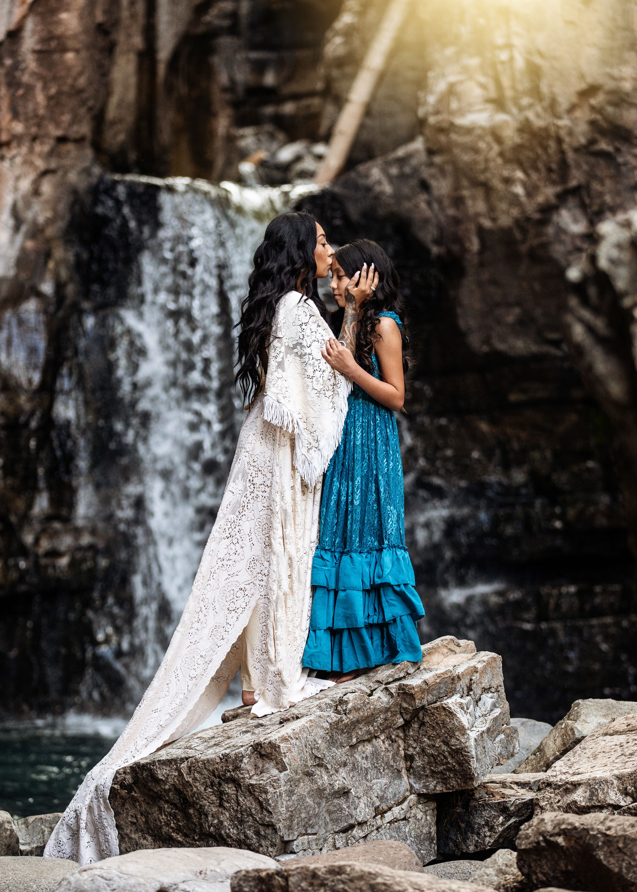 Two women sharing a kiss on rocks before a cascading waterfall in sunlight.