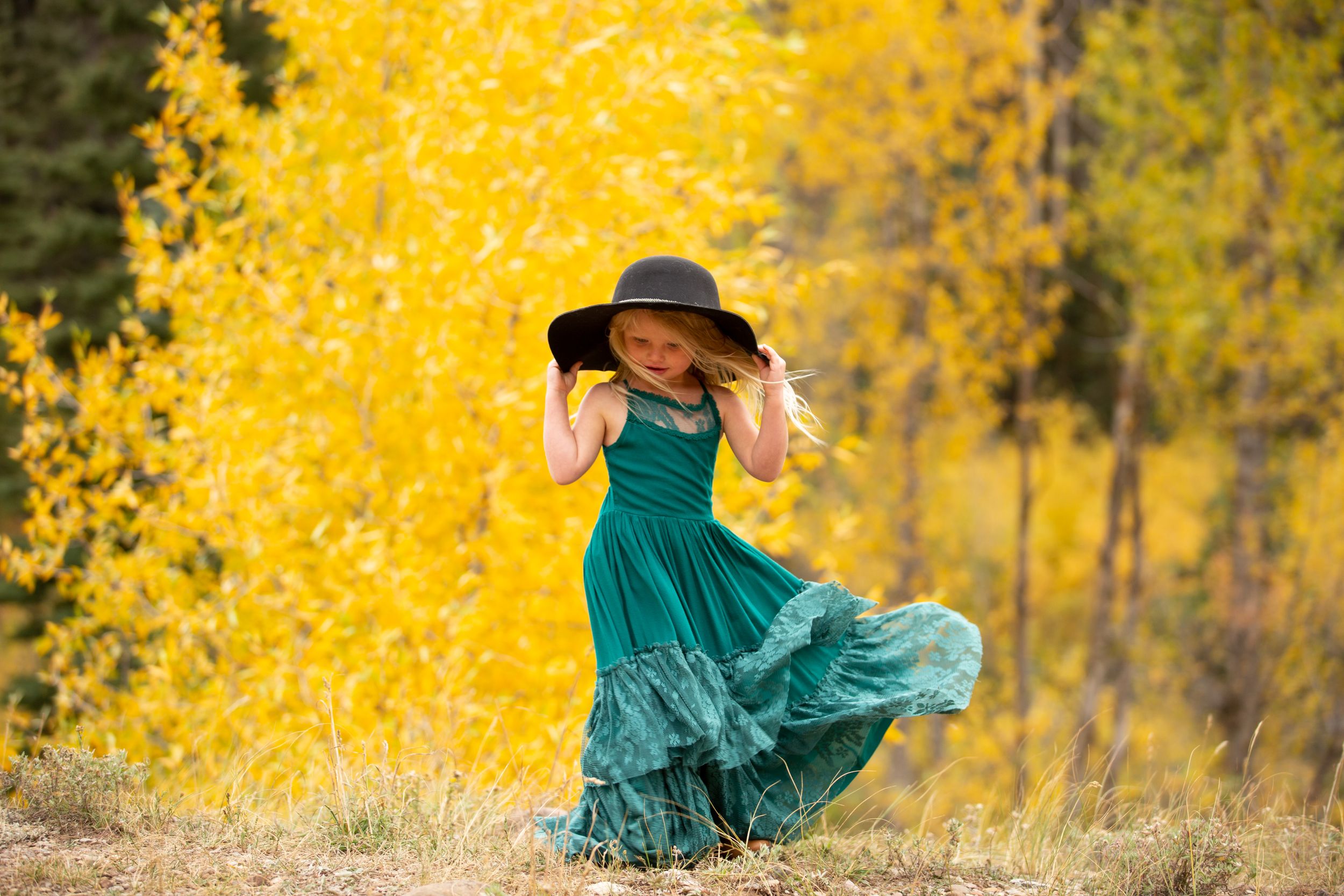 Woman in teal dress and black hat twirling among vibrant yellow autumn aspen trees.