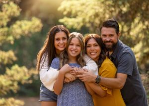 Happy family of four embracing outdoors with warm sunlit green trees behind them.