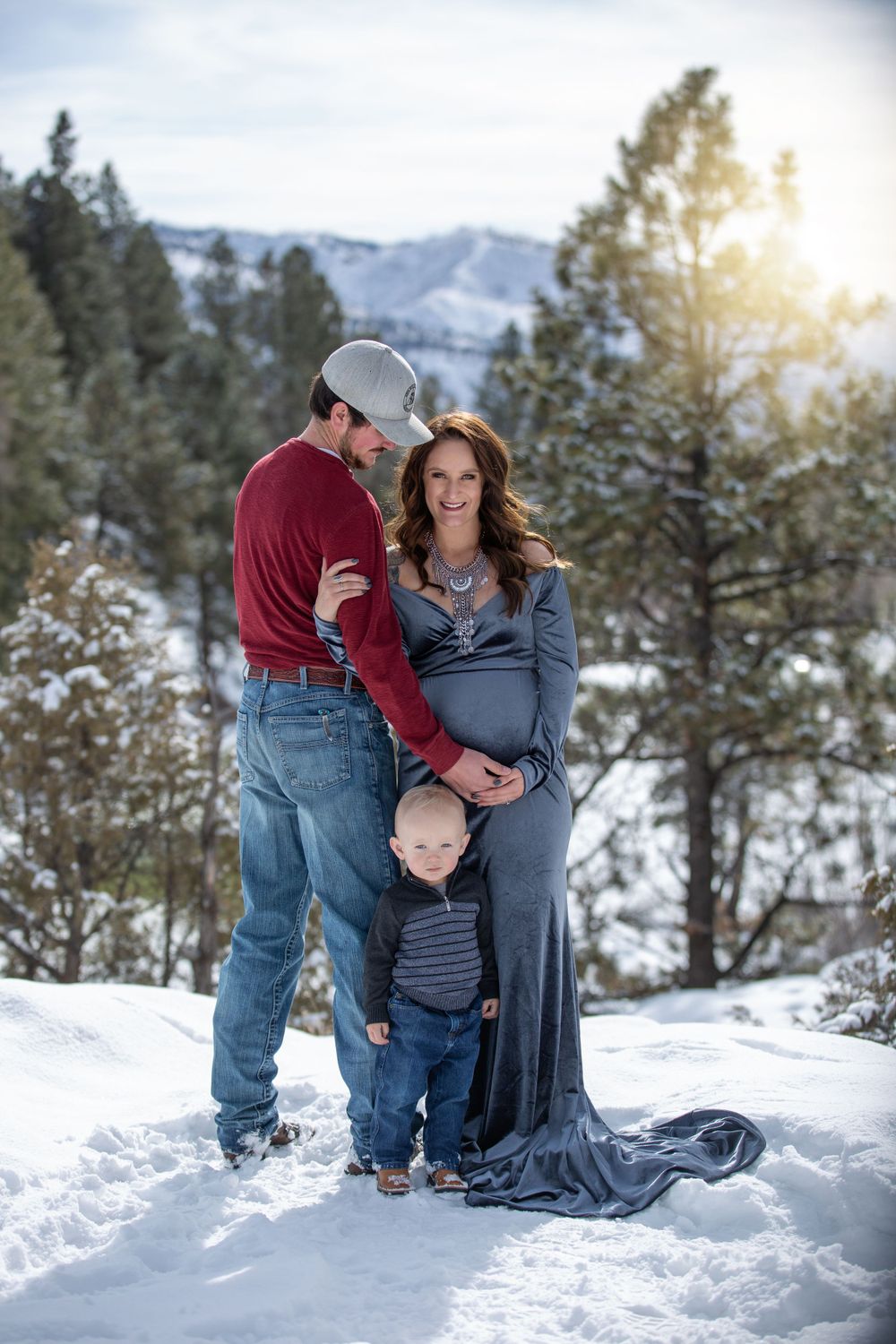 Maternity photo of pregnant couple with toddler in snowy mountain landscape at sunset.
