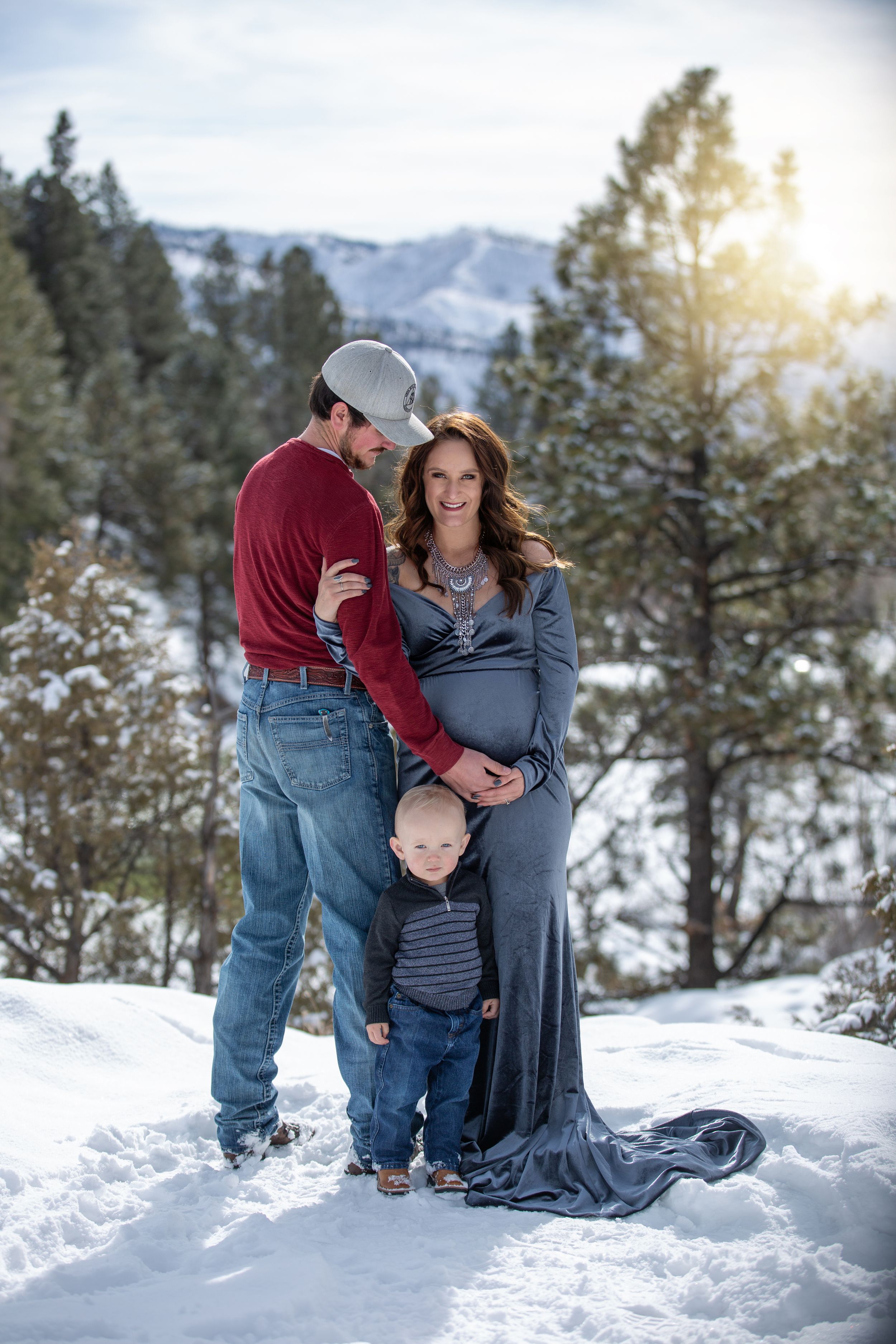 Maternity photo of pregnant couple with toddler in snowy mountain landscape at sunset.