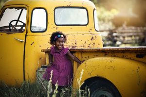 Joyful young girl in purple dress leaning against a vintage yellow truck outdoors.