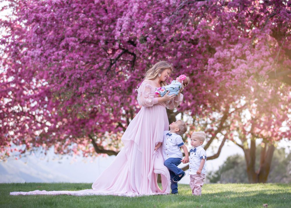 Mother in pink gown holding newborn under blooming pink tree with toddler beside her in spring.
