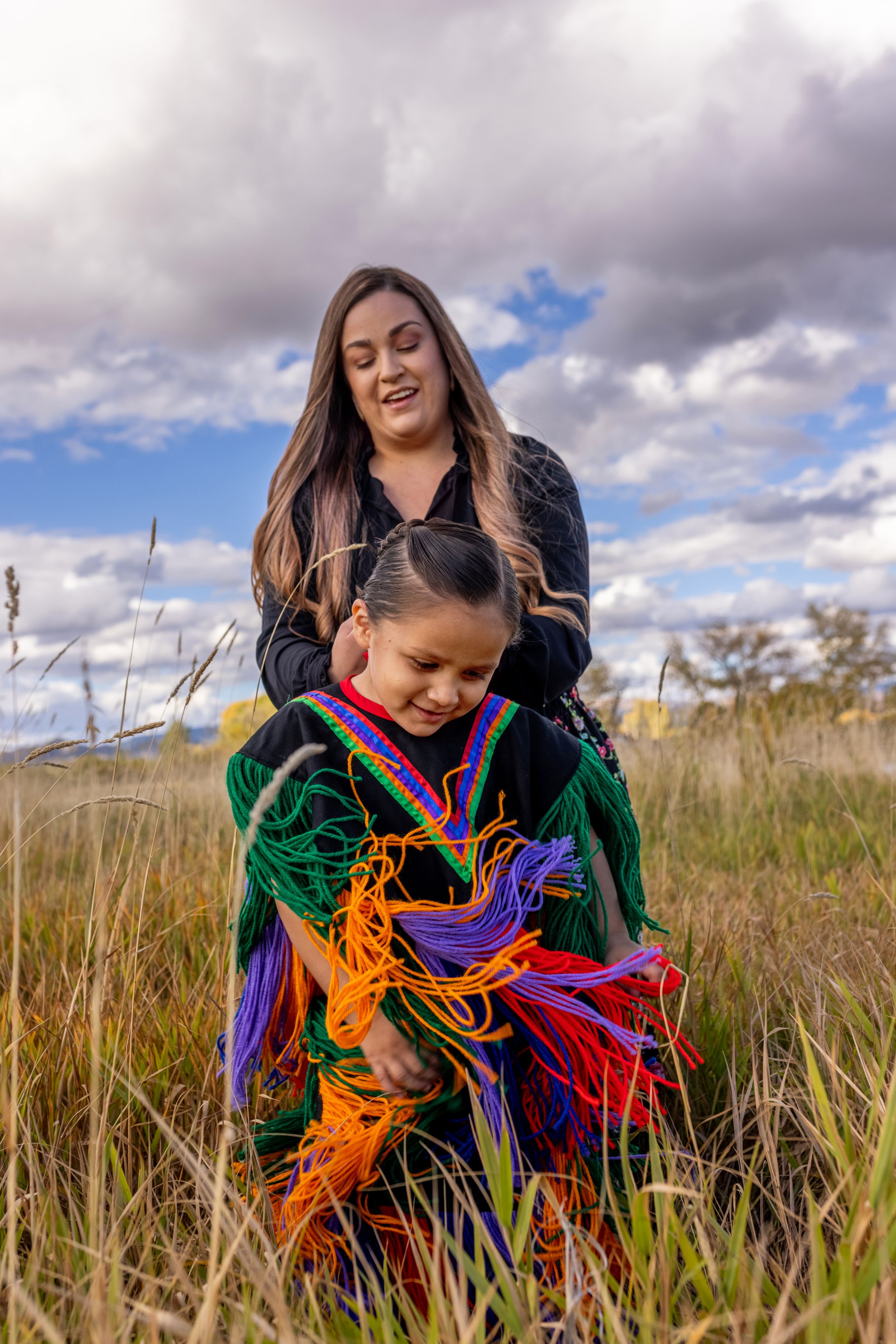 Native American mother braiding daughters hair in colorful traditional regalia in golden meadow.