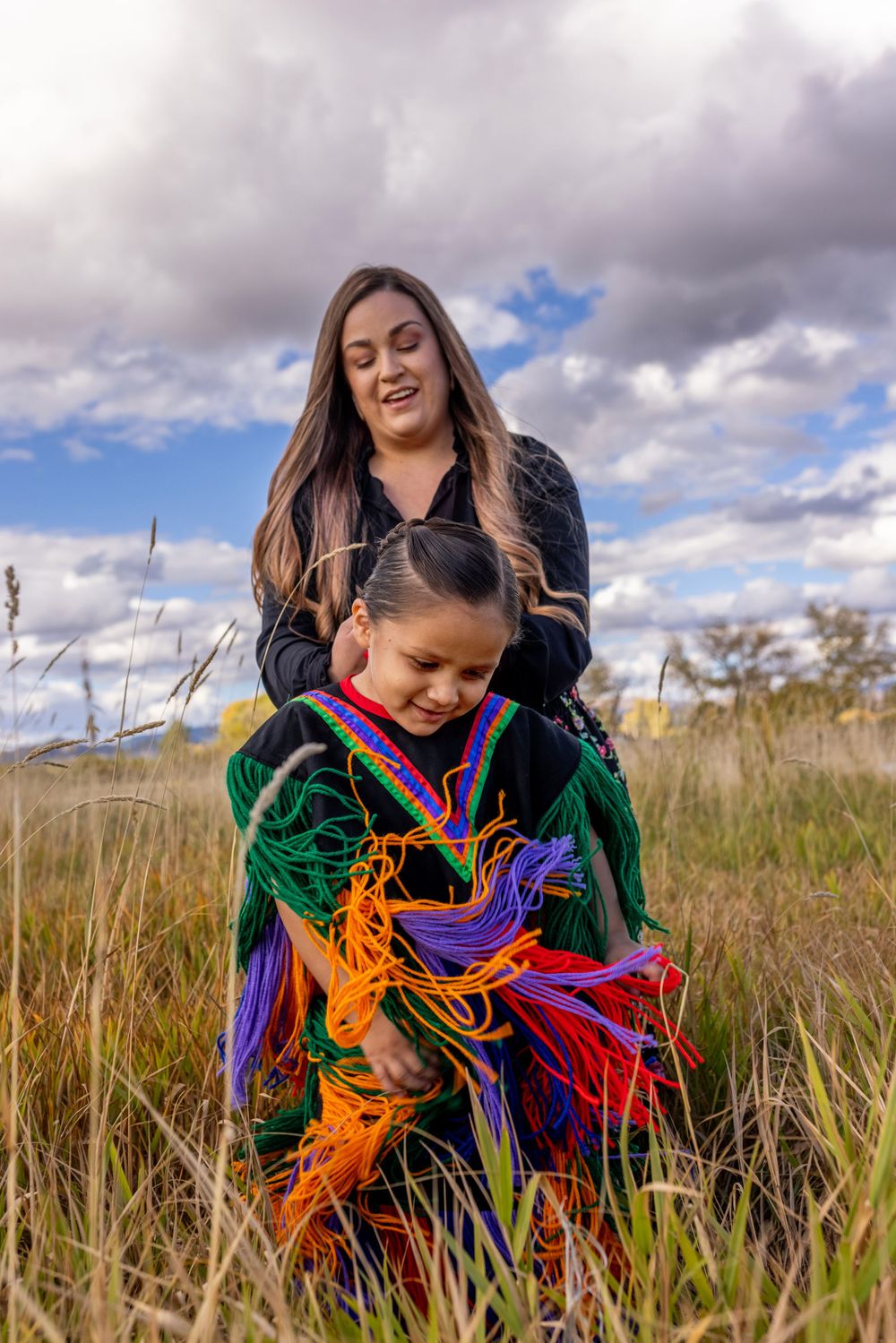 Native American mother braiding daughters hair in colorful traditional regalia in golden meadow.