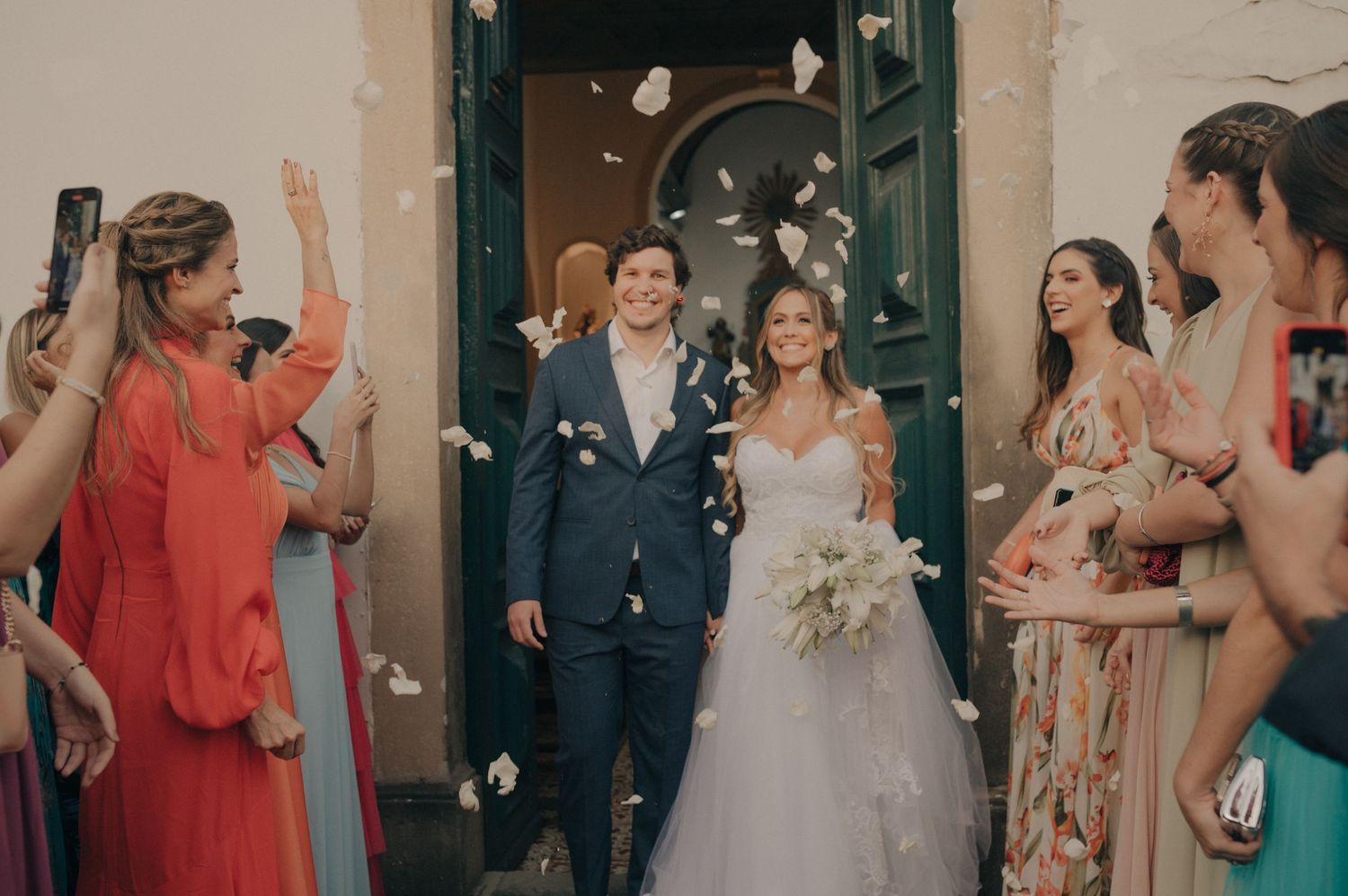 Happy newlyweds exit their ceremony while guests throw petals and celebrate with raised hands.