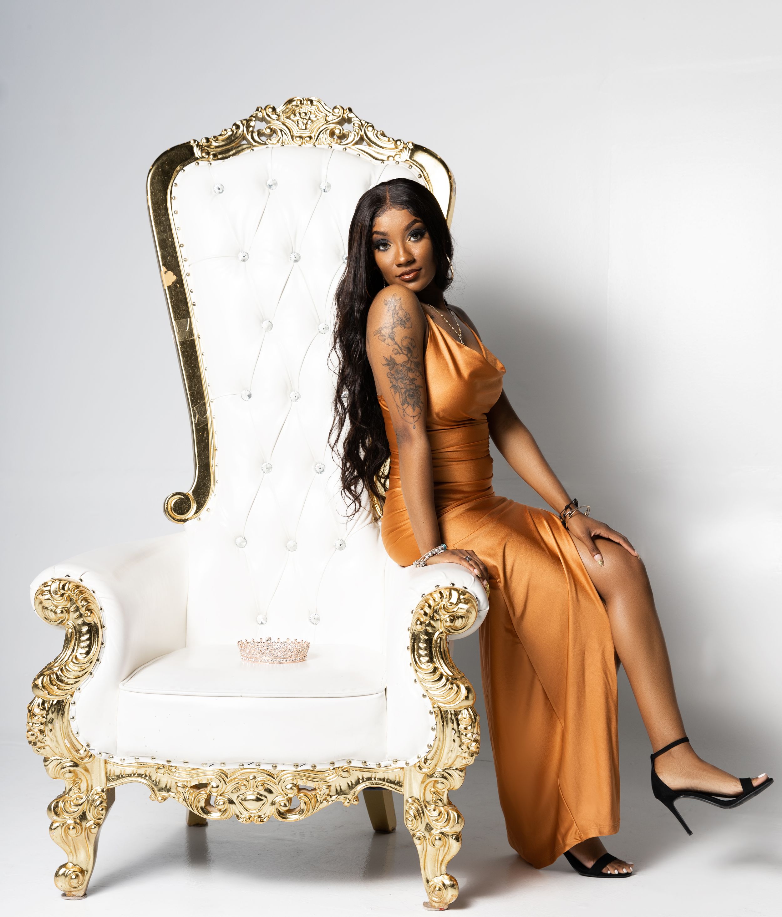 Model in orange gown poses on ornate white and gold throne chair against white background.
