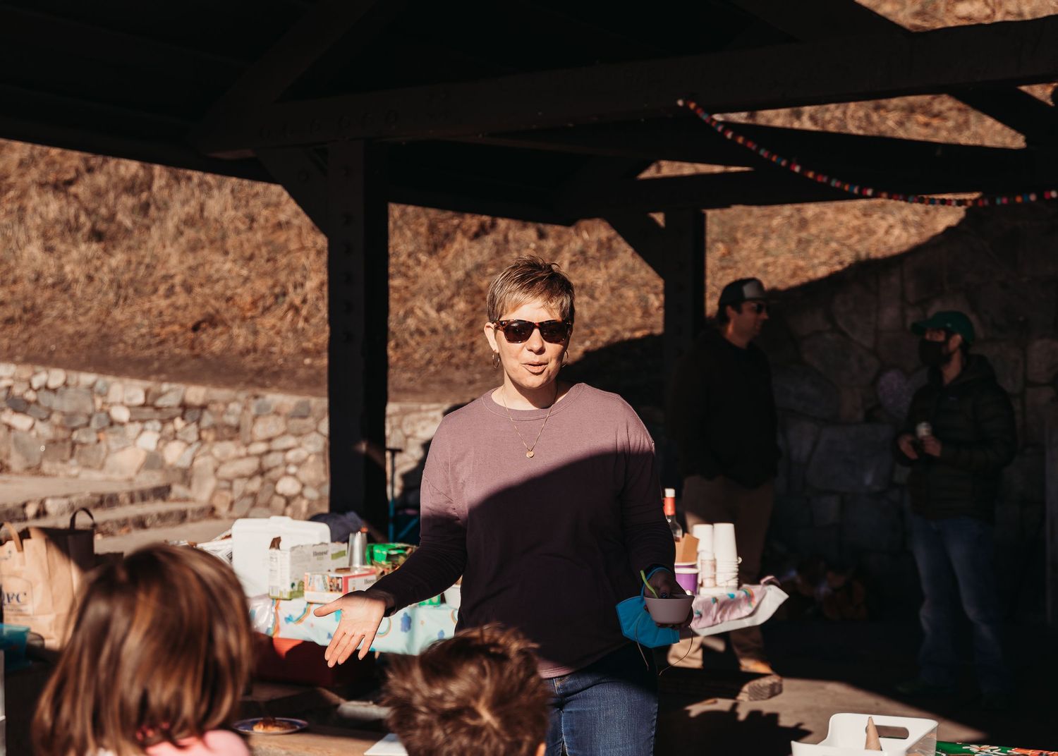 Person in sunglasses at outdoor gathering serving food under covered shelter.