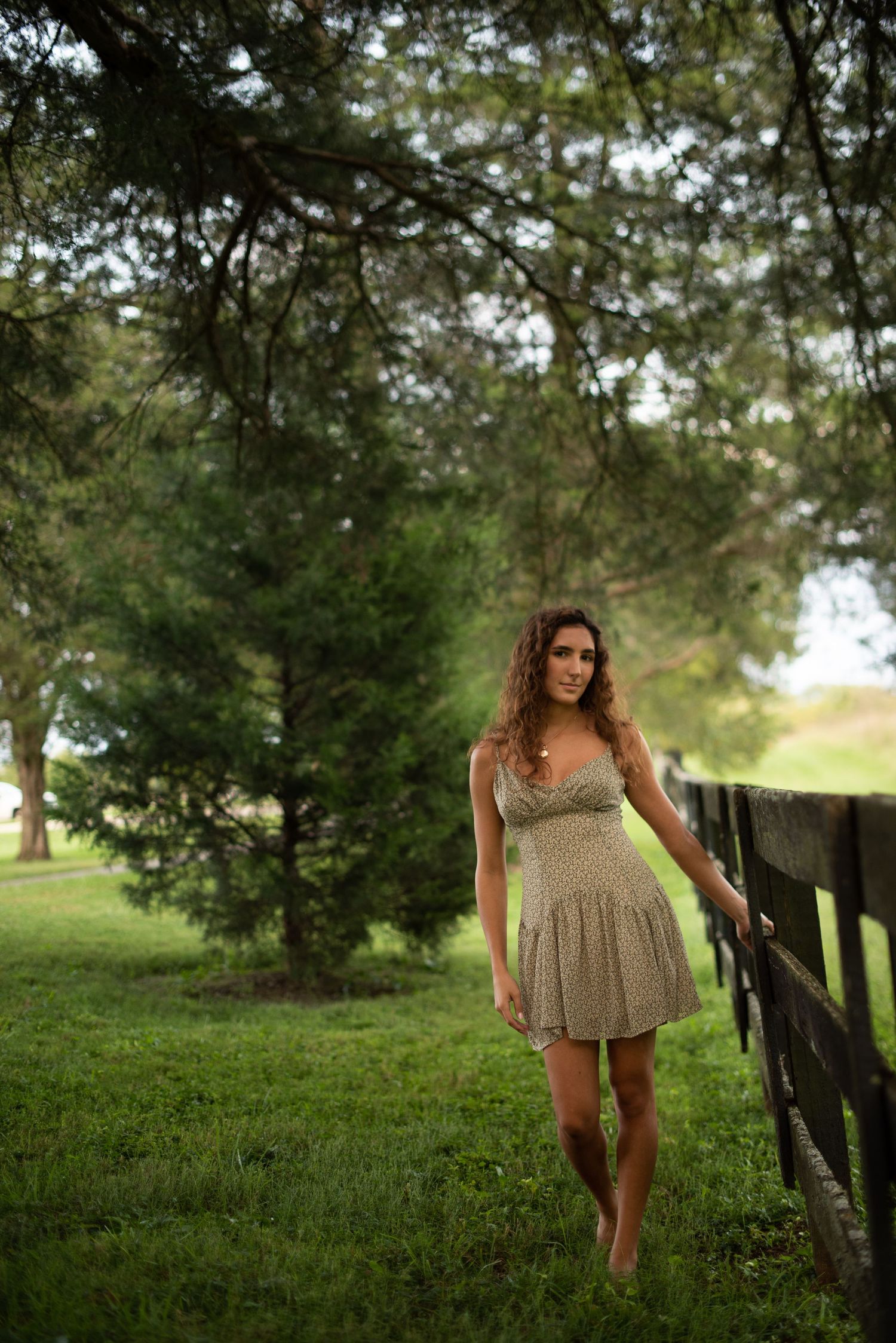 Barefoot figure in a light dress poses by a rustic wooden fence in a lush green countryside setting.