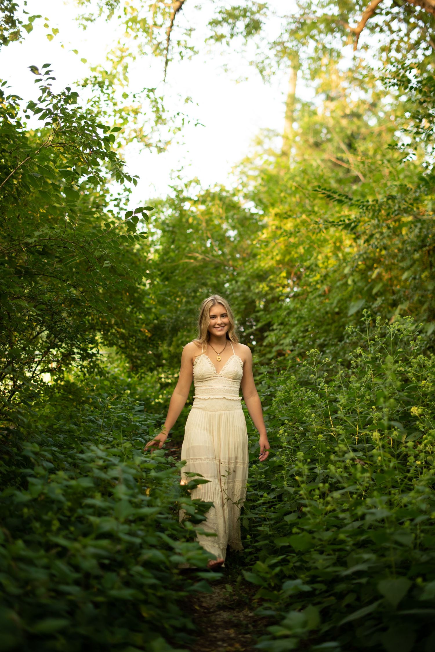 Person in flowing cream dress standing on nature path surrounded by green foliage.