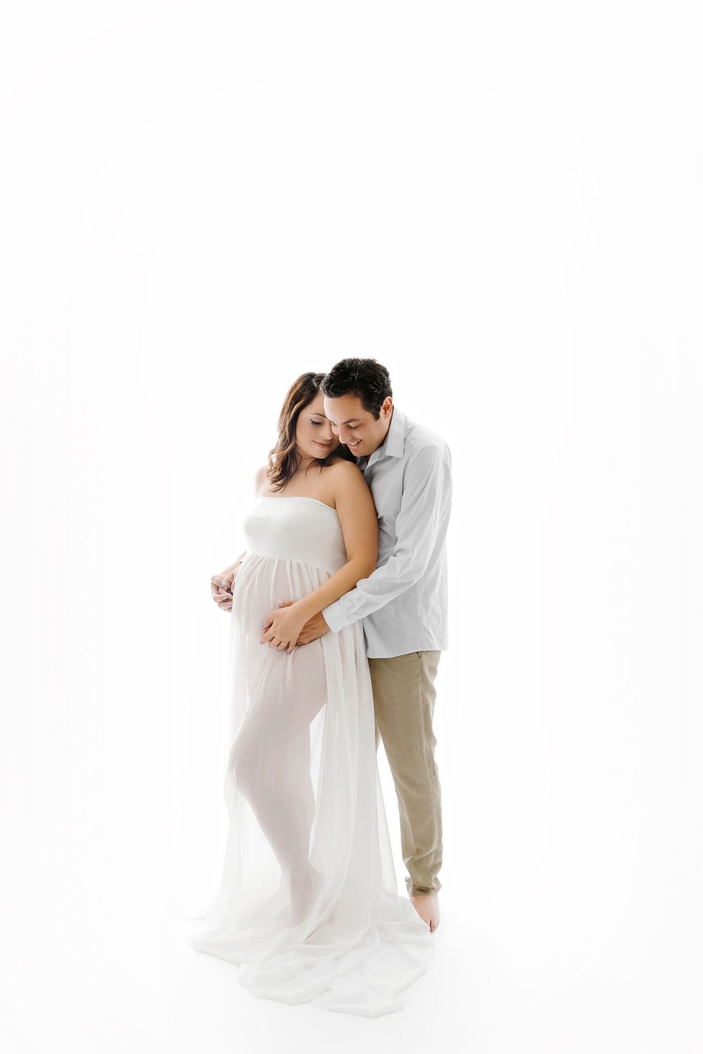 Intimate maternity portrait of couple embracing in white flowing dress against bright background.