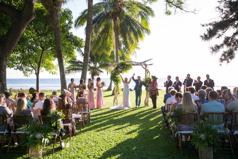 Outdoor wedding ceremony under palm trees on a lush green lawn overlooking the ocean in Hawaii.