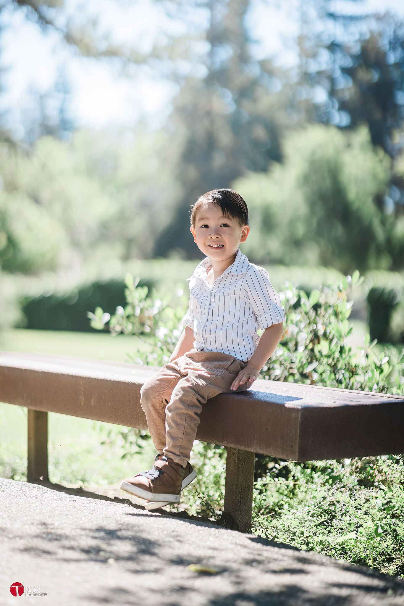 A child sits alone on a wooden bench in a sun-dappled garden path.