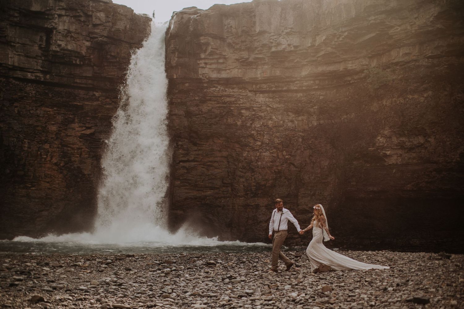Two figures stand beneath a majestic waterfall cascading down towering rock cliffs.