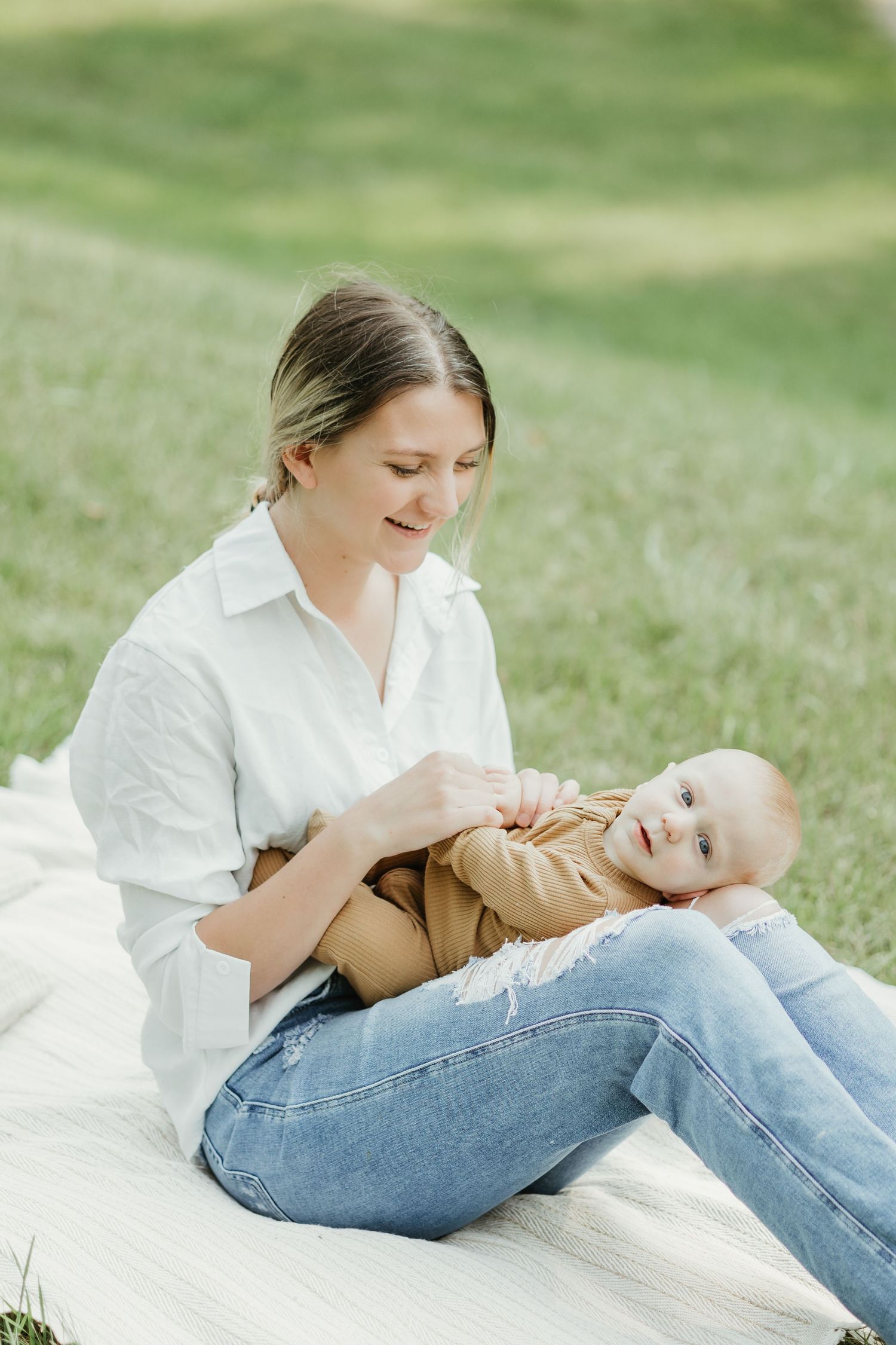 Mother and baby sharing tender moment on white blanket in grassy meadow.
