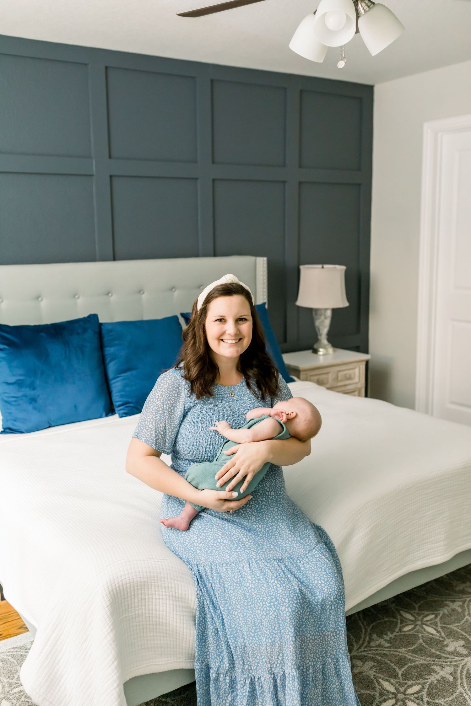 A tender moment in a bedroom with soft textures and a sage green accent wall features a light blue dress and newborn.
