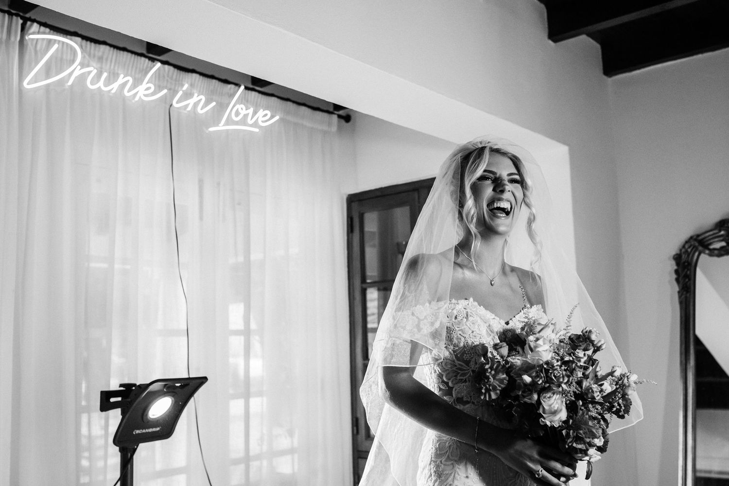 Black and white photo of a bride in wedding dress holding bouquet with neon sign reading Drunk in love behind her at Quinta da Bichinha