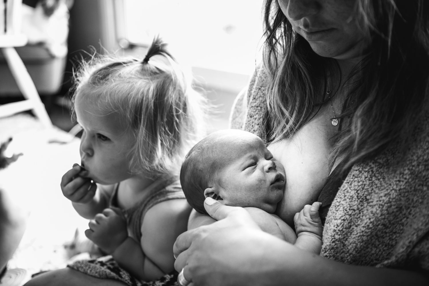 A newborn lays with his head against his mother's breast, his sister can be seen in the background.