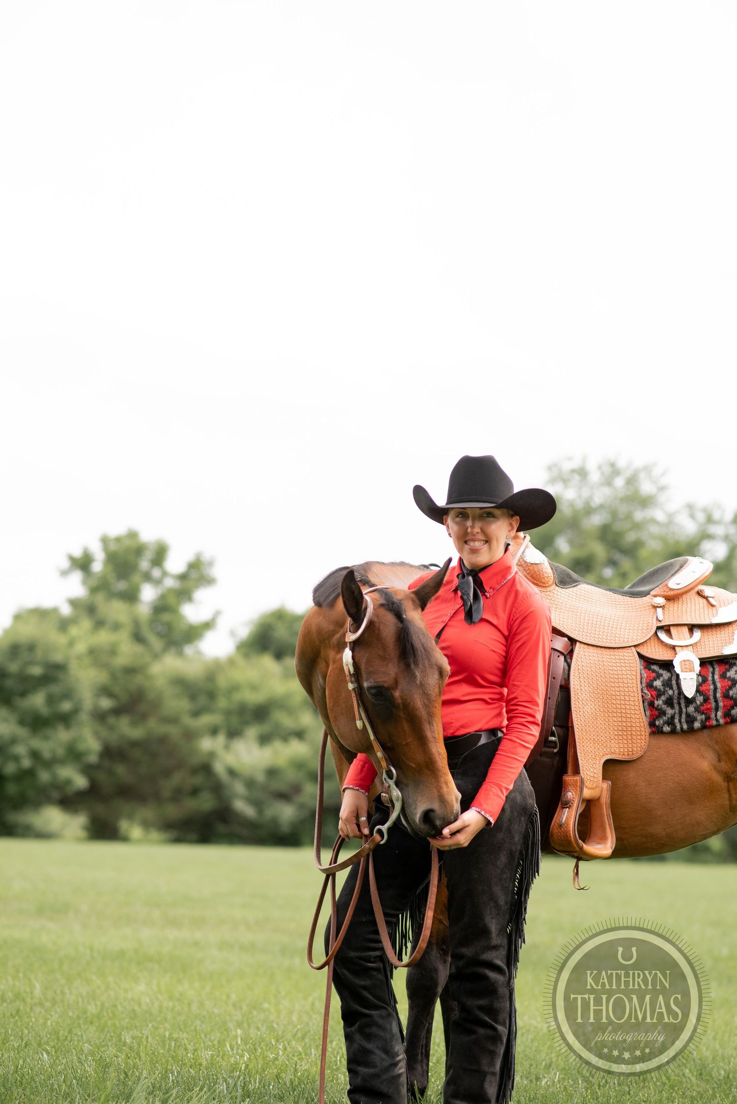 Handler in red western shirt and cowboy hat stands with brown horse wearing decorated saddle.