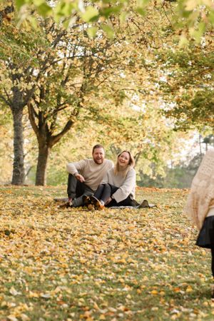 Couple sitting on autumn ground surrounded by fallen yellow leaves and trees.